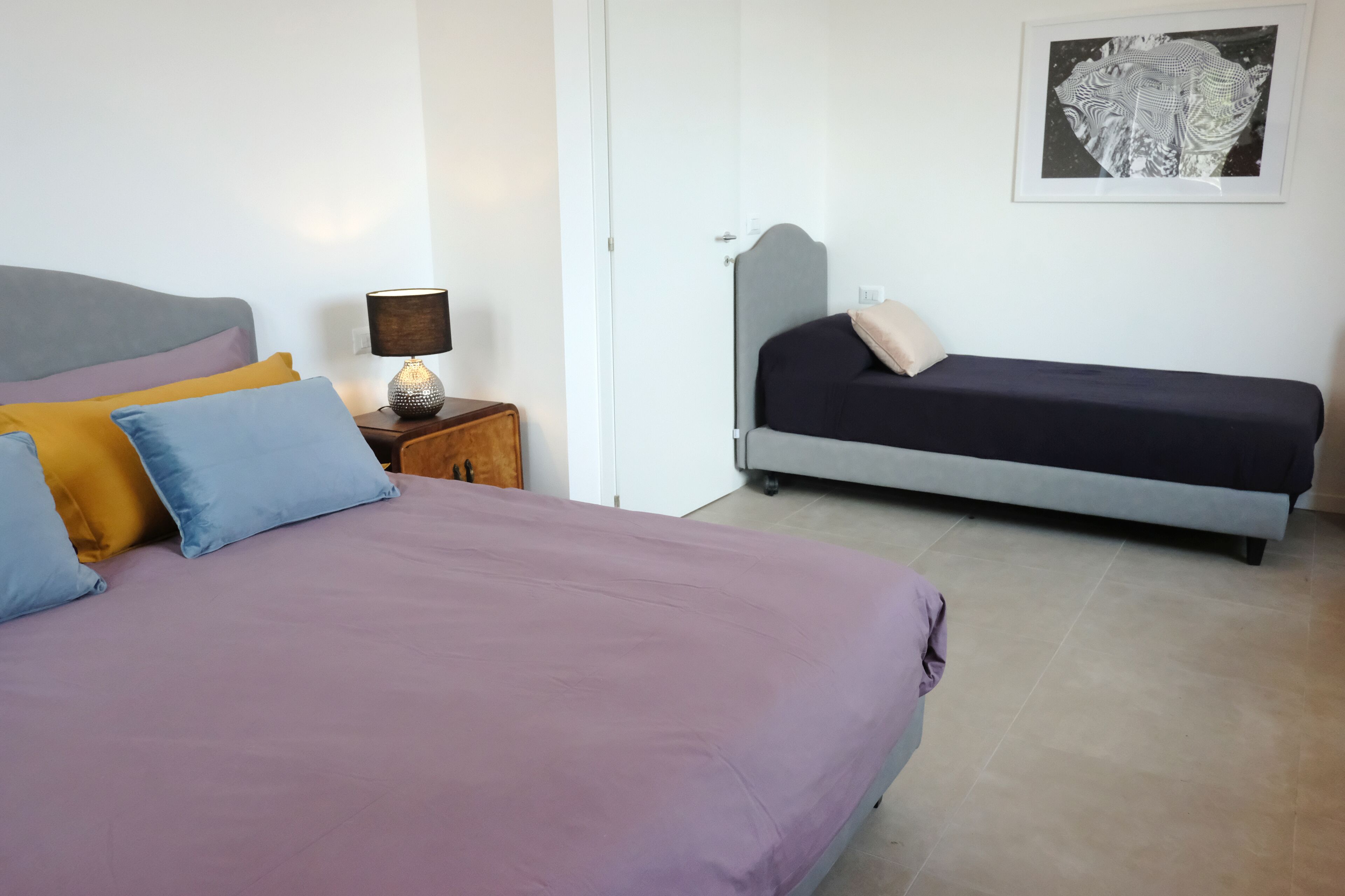 Ein Schlafzimmer mit einem großen Bett, einem kleineren Bett, einem Nachttisch mit Lampe und einem gerahmten Bild an der Wand.