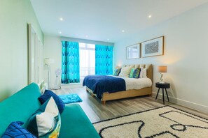 Une chambre moderne avec un lit, un canapé, un ventilateur sur pied et une table de chevet avec une lampe.
