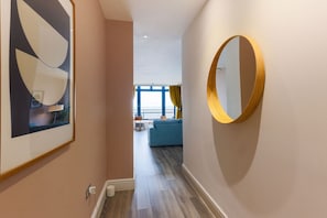 Um corredor com uma obra de arte abstrata emoldurada, um espelho redondo e vista para uma sala de estar com um sofá azul e cortinas amarelas.