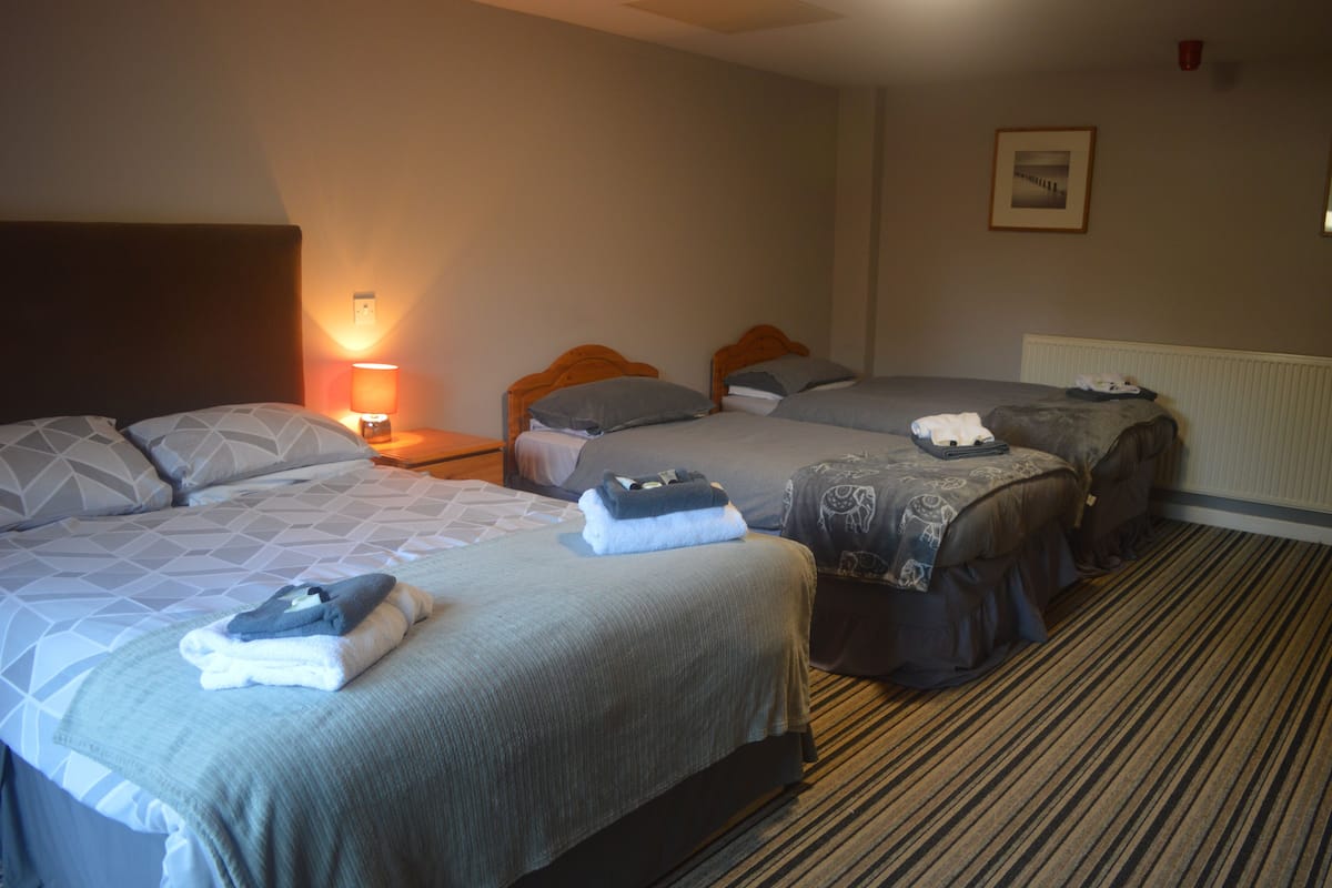 A hotel room with three beds, each with a different headboard design. There is a lamp on the left side, a framed picture on the wall, and a radiator near the window.