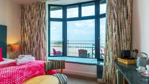 A hotel room with a large bay window, a bed with pink bedding, a small round table, a chair, a desk with a kettle, and a view of the sea.