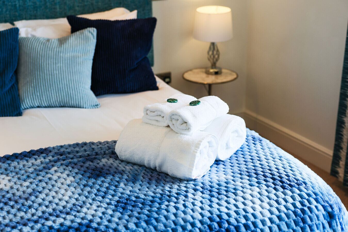 A neatly made bed with a blue textured bedspread, a stack of white towels with green tags, and a bedside lamp on a round table.