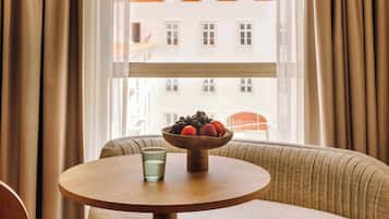 Una mesa redonda de madera con un vaso de agua y un cuenco de frutas de madera frente a una ventana con cortinas.