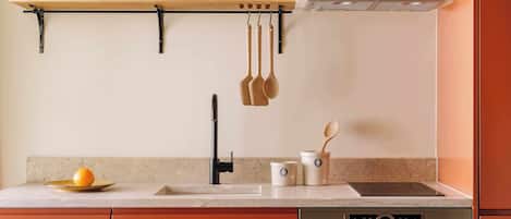 A kitchen with a wooden bowl of oranges on a shelf, a hanging pot rack, a sink, a stove, and a cabinet.