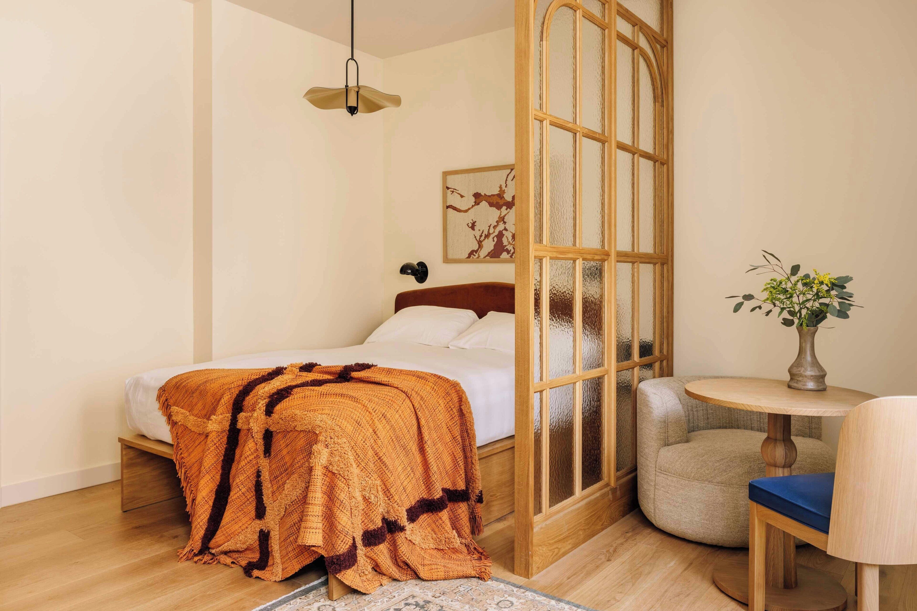 A bedroom with a bed, a round wooden table, a chair, a vase with flowers, and a wall-mounted artwork.