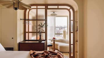Habitación de hotel con cama, cómoda, una mesita con fruta, una silla y vistas a la ciudad a través de una ventana.