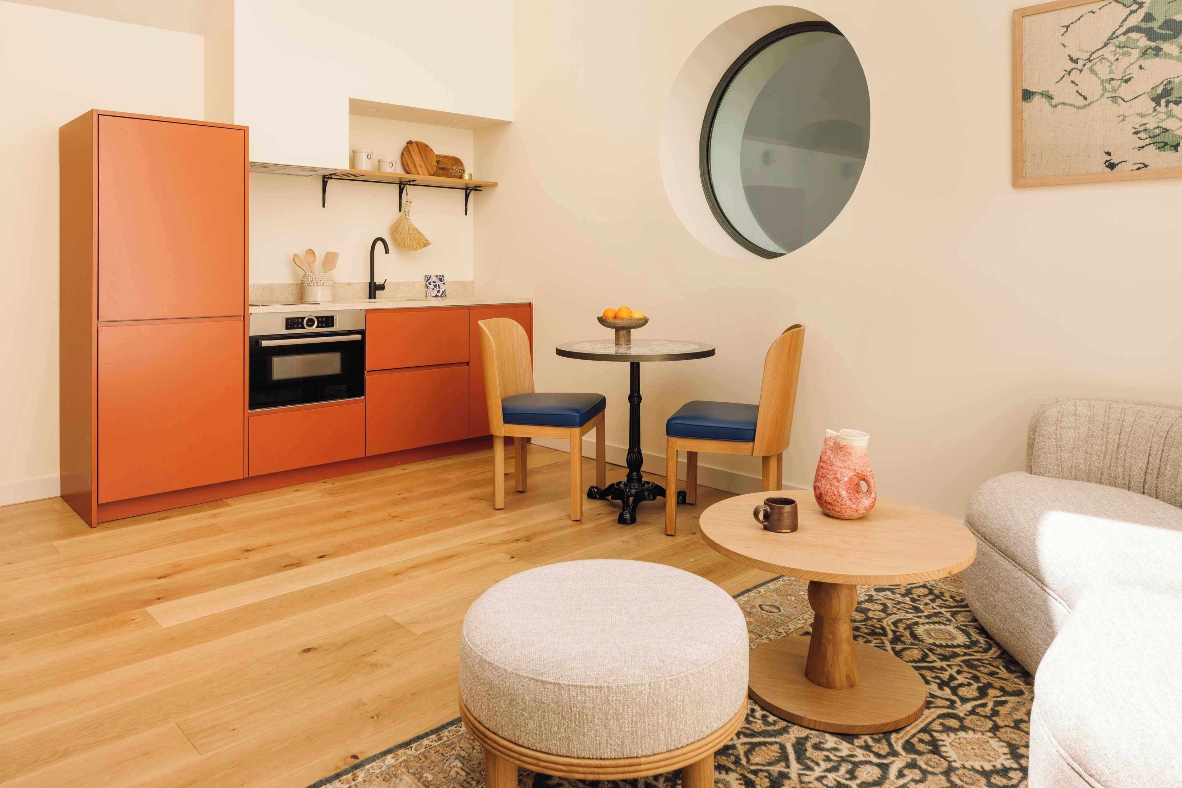 Un espacio habitable compacto con kitchenette, una mesa de comedor redonda, dos sillas, un reposapiés redondo y un sofá.