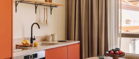 A compact kitchen with a built-in oven, a sink, and a wooden shelf above it. There's a bowl of fruit on a small table next to the window.