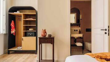 Un dormitorio con cama, una mesita de noche con un florero, un armario de madera y un baño visible a través de una puerta abierta.