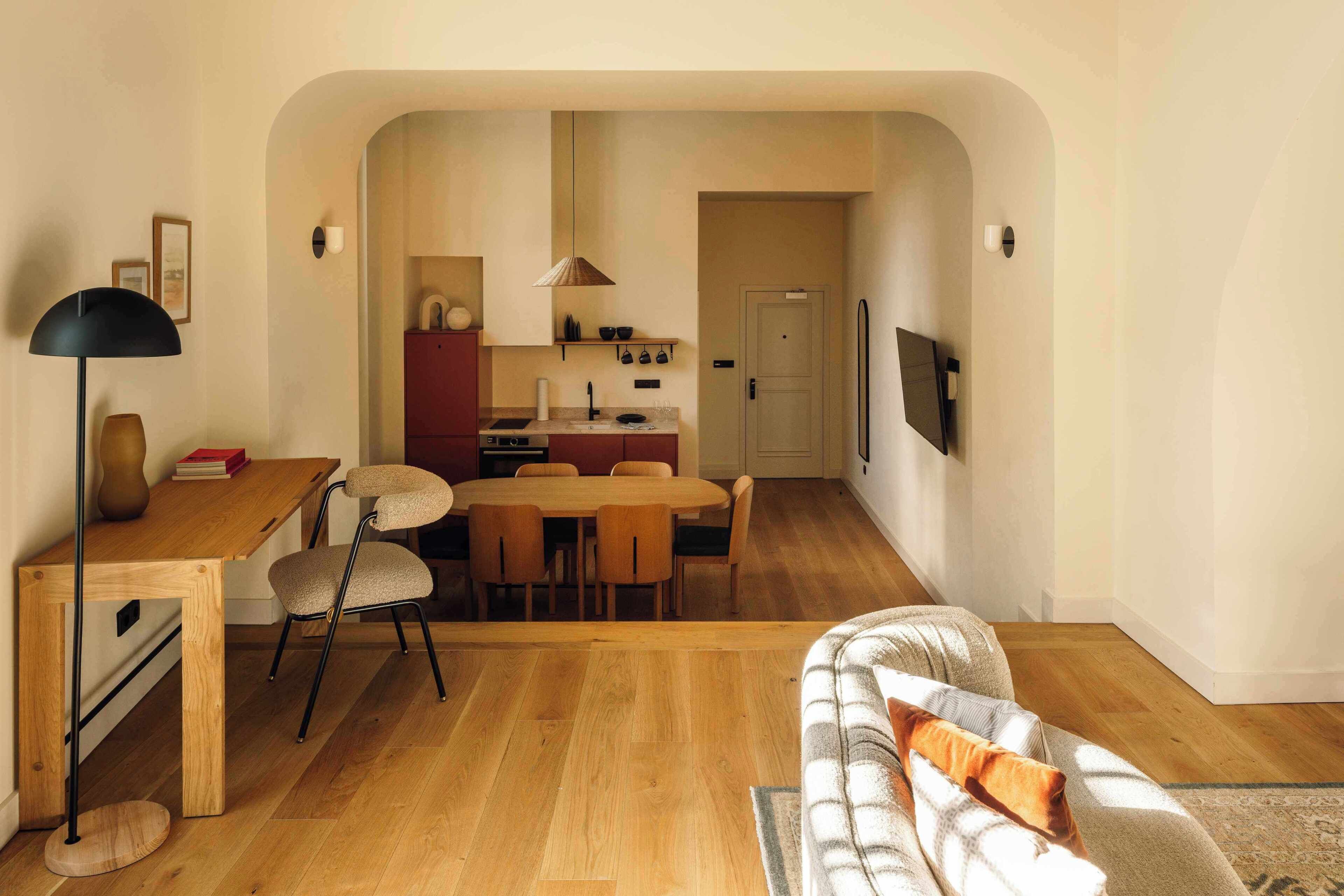 A modern living room with a wooden floor, a sofa, a dining table with chairs, a kitchen area with a stove and cabinets, and a TV mounted on the wall.