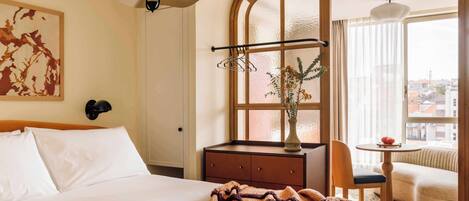 A hotel room with a bed, a desk with a chair, a window with a view, and a hanging light fixture.