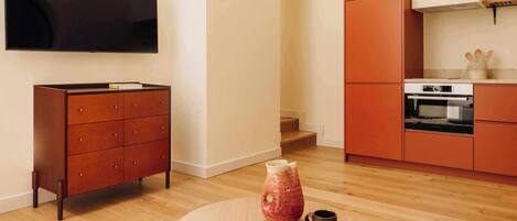A modern living room with a flat-screen TV, a wooden cabinet, a kitchen area with orange cabinets, and a round coffee table with a vase and mug.