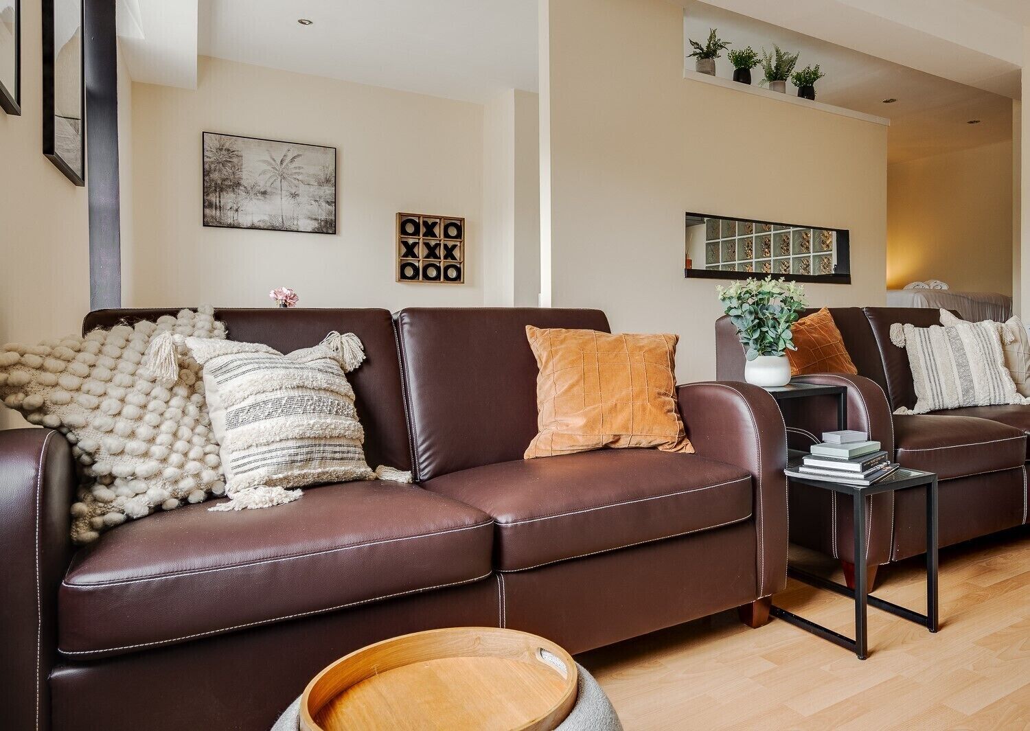 A brown leather sofa with multiple cushions, a small wooden side table with a plant, and a framed artwork on the wall.