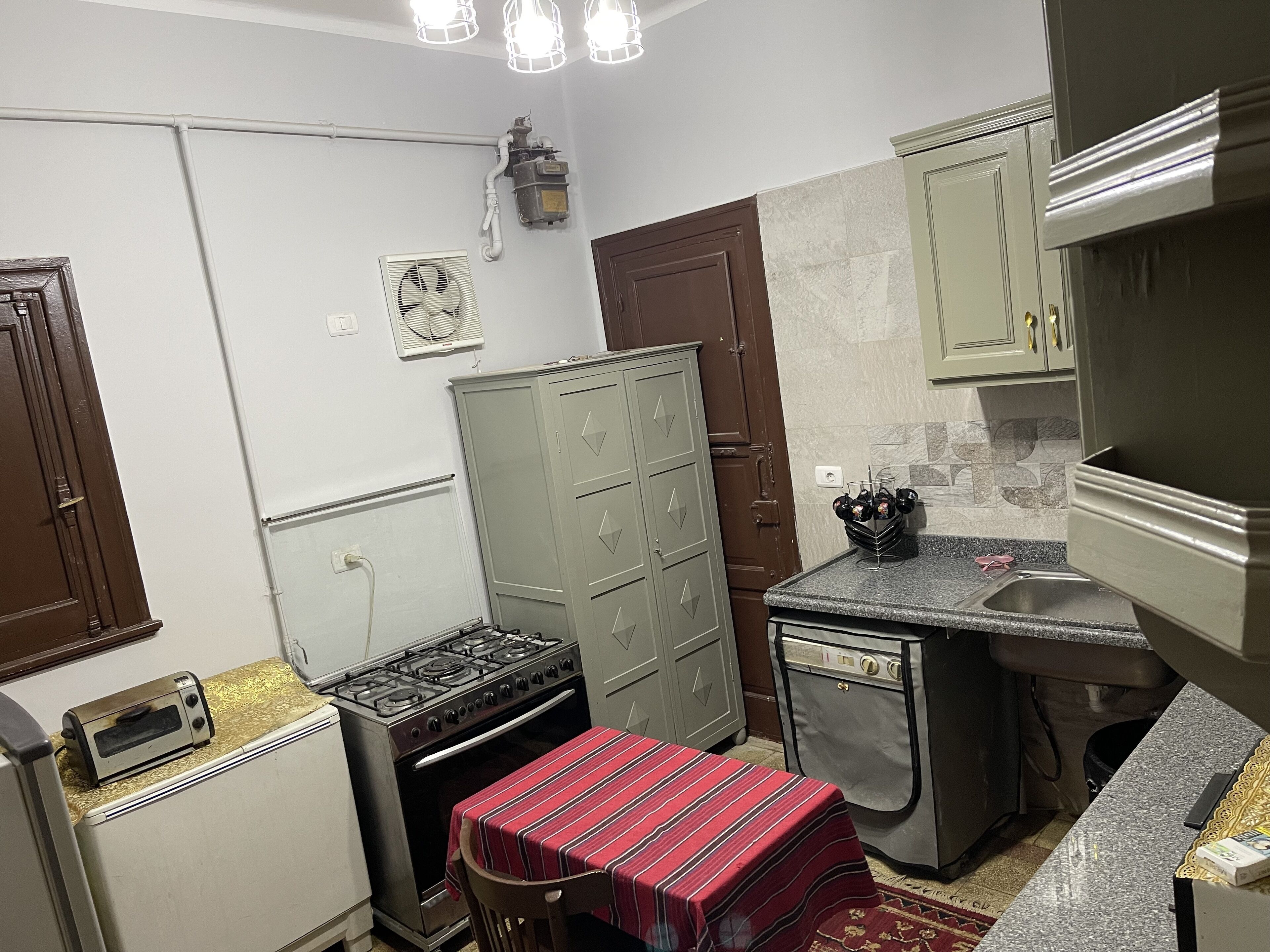 Una cocina compacta con estufa a gas, horno pequeño, pileta y una mesa con mantel a rayas.
