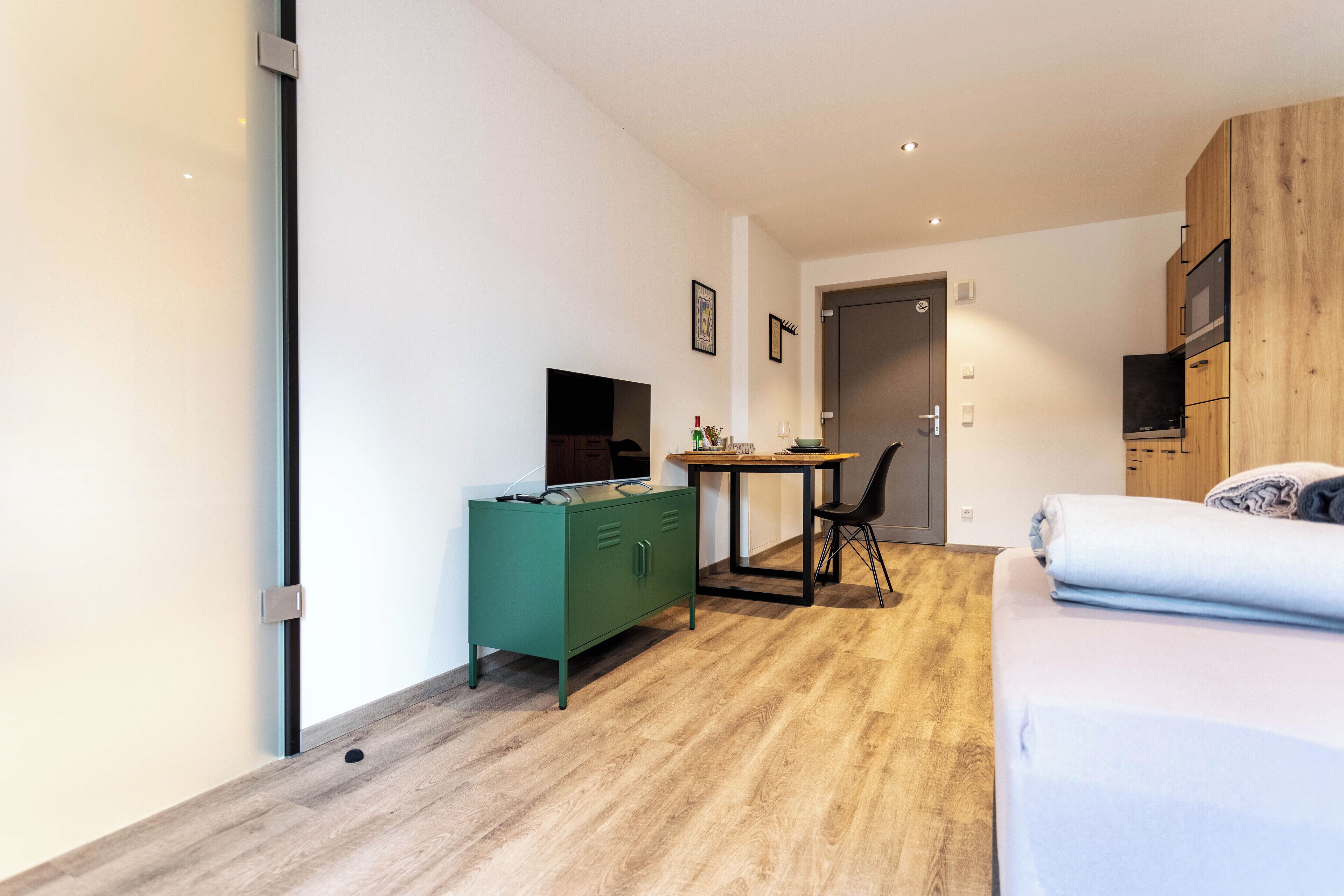 A modern hotel room with a wooden floor, a green minibar, a desk with a chair, a bed, and a TV mounted on the wall.