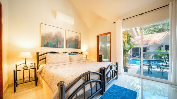 A bedroom with a bed, bedside tables, a window with a view of a pool and outdoor furniture, and artwork on the wall. - Villa Perla 15 At Playacar phase ll NAS (Playa del Carmen)