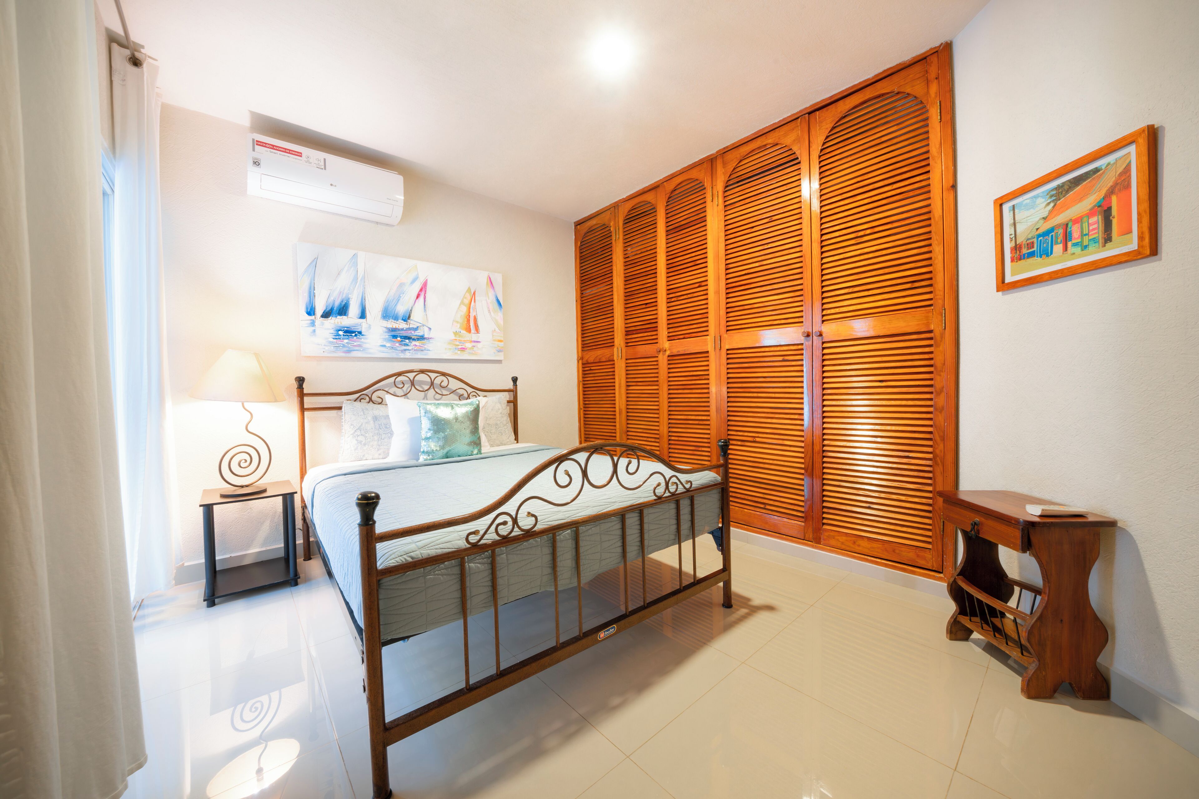 A bedroom with a bed, wooden wardrobe, a nightstand, a painting, and an air conditioning unit.