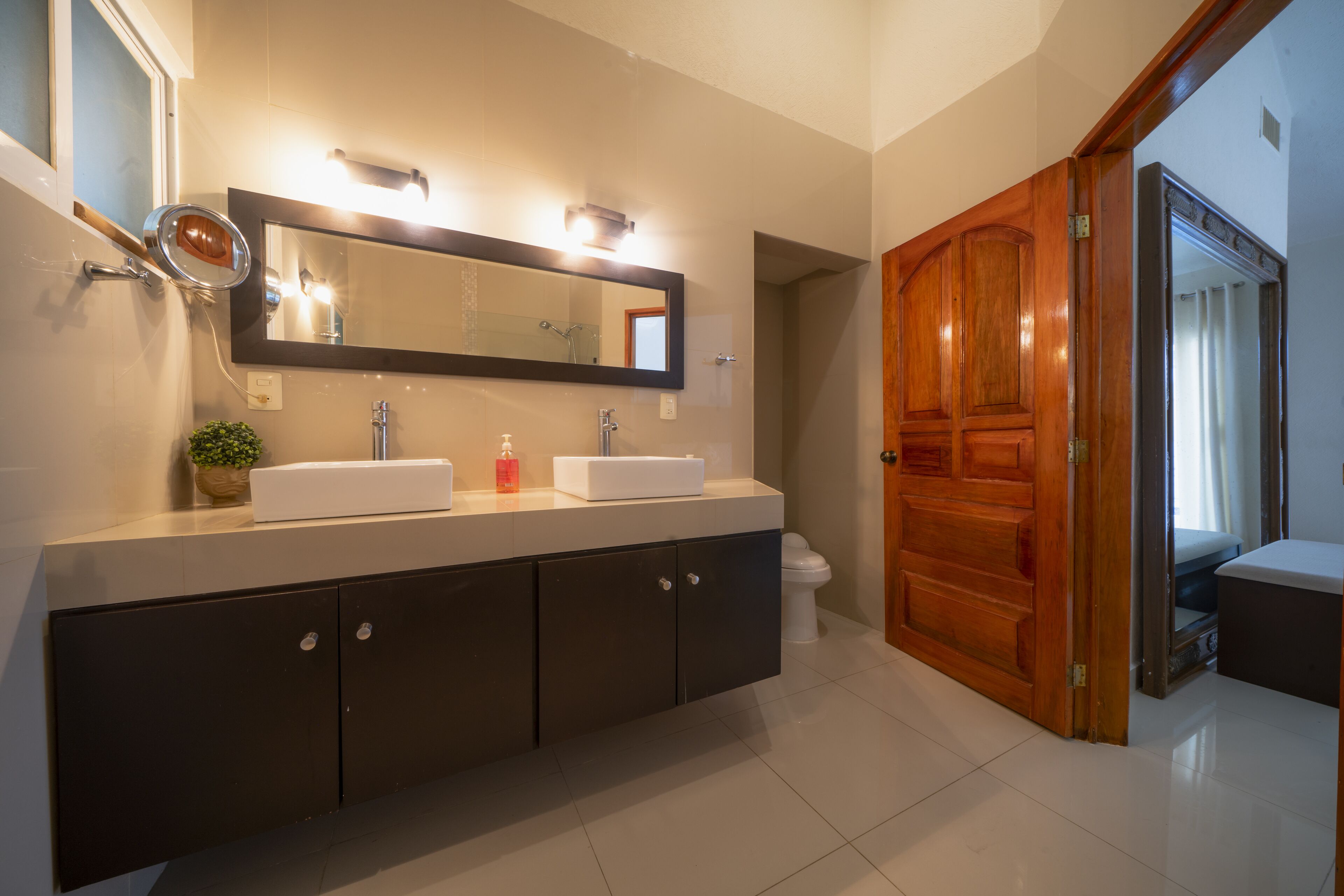 Un baño moderno con un espejo grande, dos lavamanos, armarios oscuros y una puerta de madera.