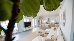 A modern living room with a white sofa, a round coffee table, and a TV. There is a kitchen in the background with white cabinets and a large window. - Sea View Villa @ Flamenco Beach, Culebra, Puerto Rico (Flamenco)