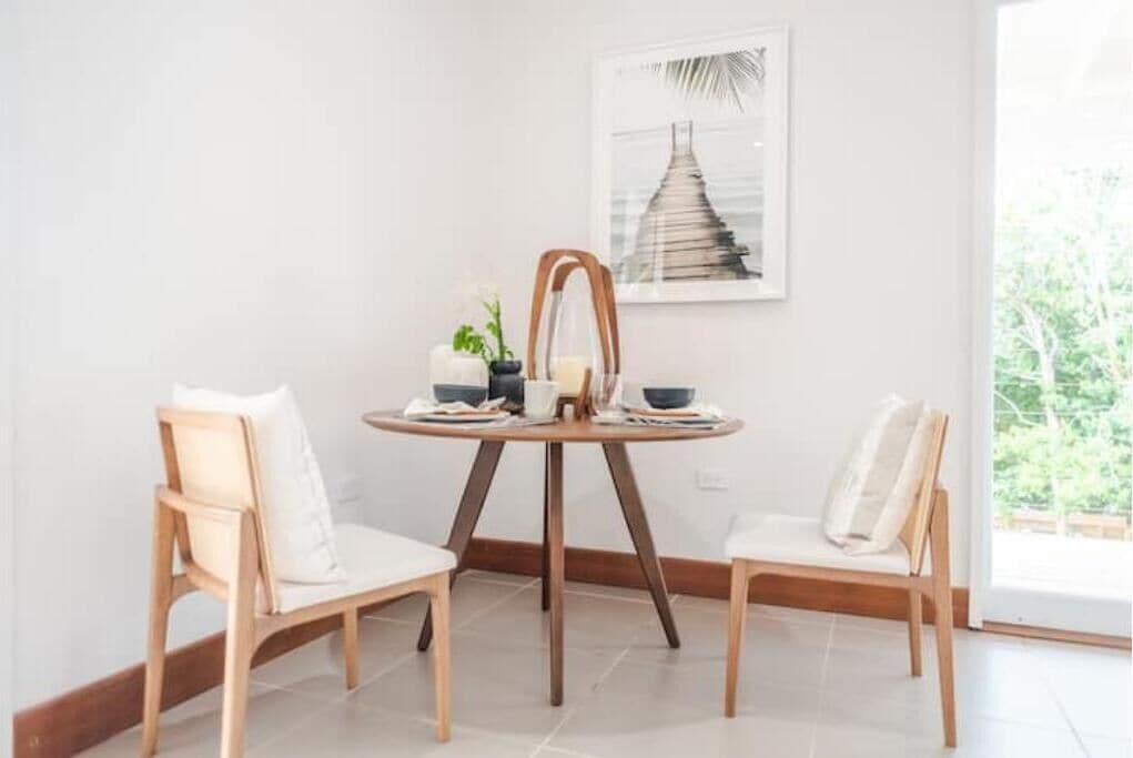 Un coin repas avec une table ronde et deux chaises, un tableau encadré au mur et une fenêtre donnant sur un paysage verdoyant.
