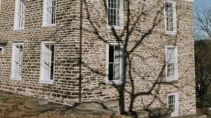 Un bâtiment en pierre doté d’une cheminée centrale, de nombreuses fenêtres et dont l’ombre est projetée par un arbre.