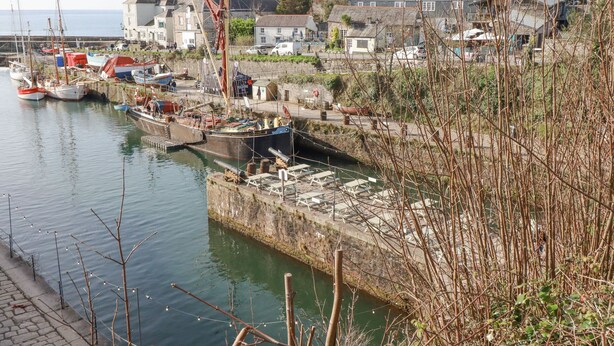 A harbor with boats docked, a stone quay, and houses in the background. - Eden Retreat (Par)
