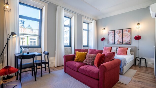 Une chambre moderne avec un canapé rouge, un lit agrémenté de plusieurs coussins, un bureau avec une chaise et de grandes fenêtres donnant sur la ville.