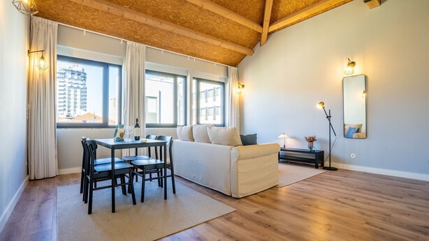 A modern living room with a wooden ceiling, a white sofa, a dining table with chairs, and a large window with curtains. - Sta. Catarina 632 - 3 by LovelyStay (Porto)
