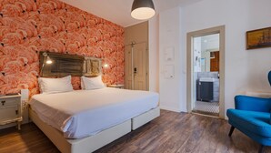 Quarto com cama, cabeceira de madeira, roupa de cama branca, uma poltrona azul e piso de madeira.