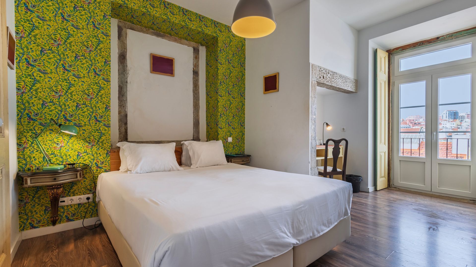 Ein Schlafzimmer mit einem großen Bett, einem Schreibtisch und einem Sessel. Ein Fenster bietet Blick auf umliegende Gebäude.