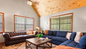 A living room with a brown leather sofa, a blue sectional sofa, and a wooden coffee table. There are large windows with blinds, a ceiling fan, and a rug with a geometric pattern. - Modern 4-bedroom Cabin w/ Hot Tub/Game Room/Fire Pit/Backyard (Sevierville)