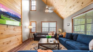 A cozy living room with a high ceiling, wooden walls, a large TV, and a blue sofa. - Modern 4-bedroom Cabin w/ Hot Tub/Game Room/Fire Pit/Backyard (Sevierville)