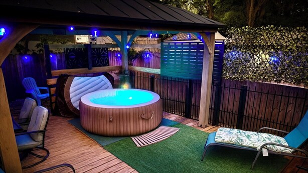 Uma banheira de hidromassagem com exterior em madeira, rodeada por um deck de madeira e grama sintética. Há cadeiras e uma mesa sob uma pérgula com iluminação azul.