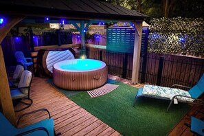 Un jacuzzi con revestimiento de madera, rodeado por una terraza de madera y césped artificial. Hay sillas y una mesa bajo una pérgola con iluminación azul.