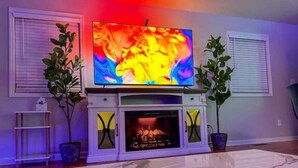 Uma sala de estar com uma televisão de tela plana em um móvel branco, lareira e duas plantas em vasos.