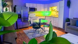 Cozinha compacta com armários brancos, micro-ondas e área de refeições com mesa e cadeiras de vidro.