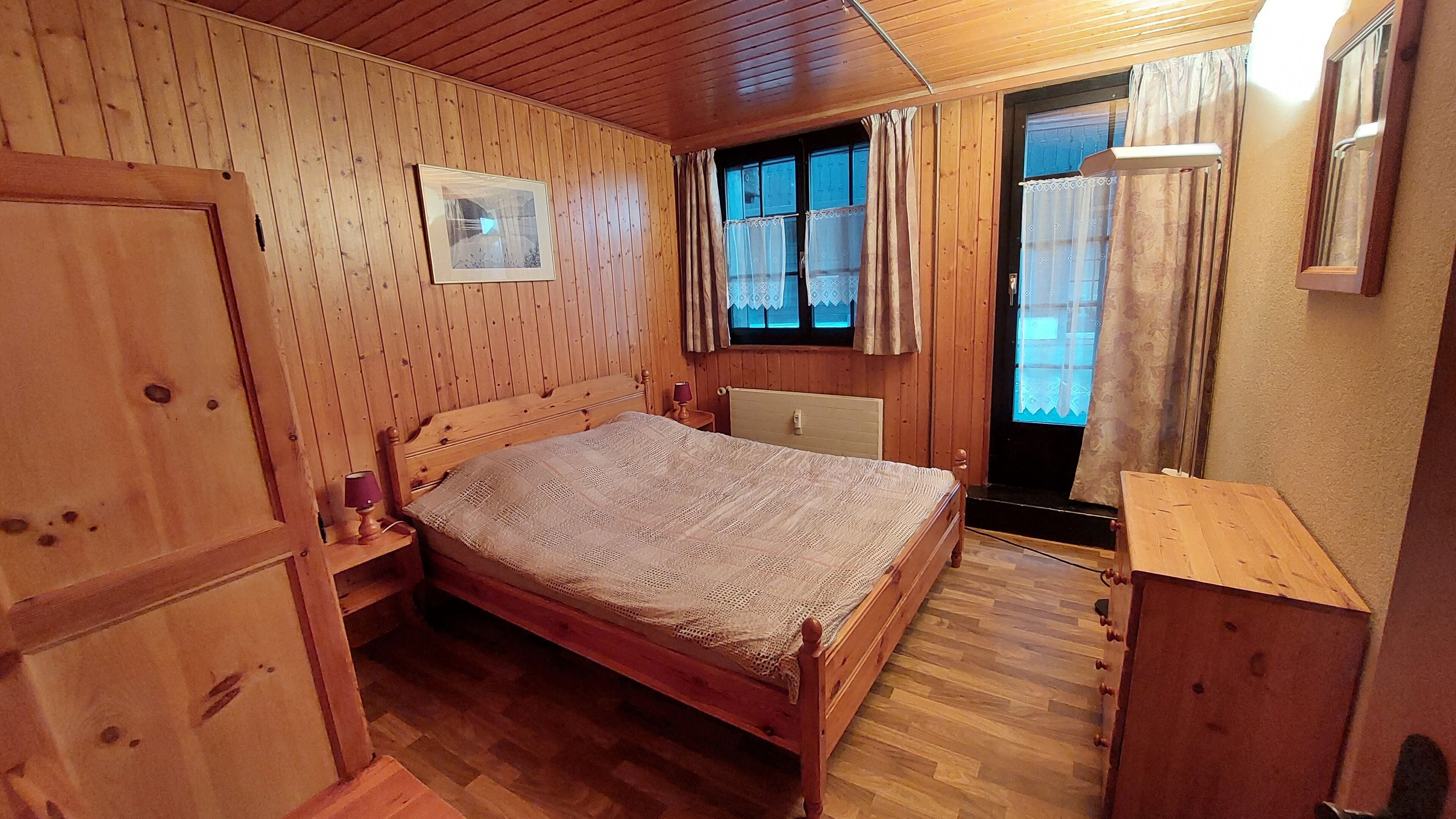 Ein Zimmer mit Holzwänden, einem Bett, einem Nachttisch, einem Fenster mit Vorhängen und einem Spiegel.