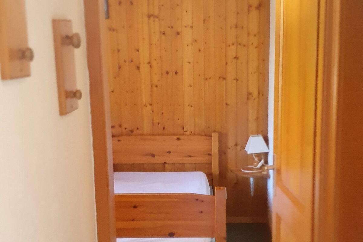 A narrow hallway leading to a bedroom with a wooden headboard, a bedside table with a lamp, and a bed with white bedding.