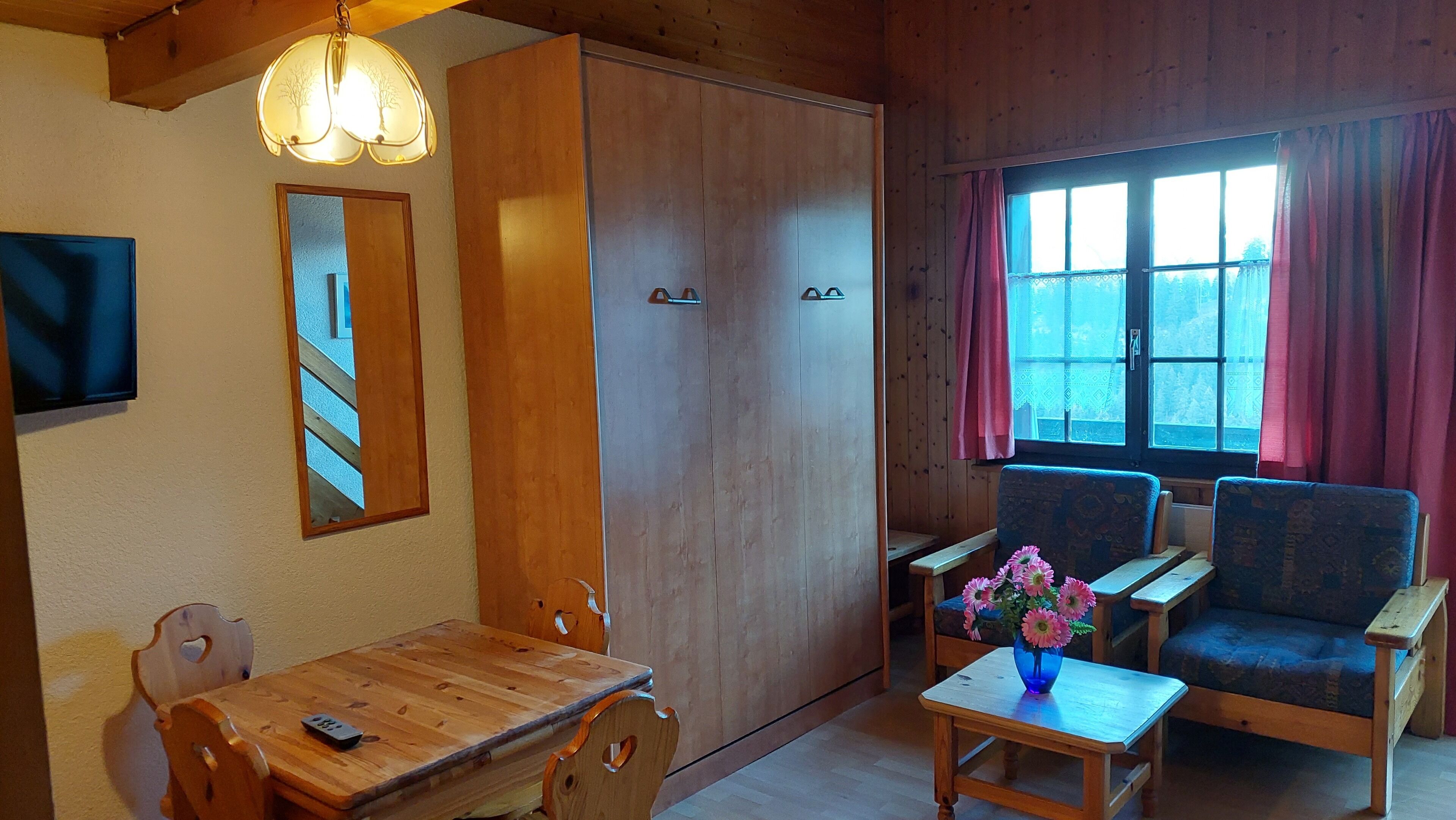A wooden interior with a dining table, chairs, a sofa, a window with curtains, and a wardrobe.