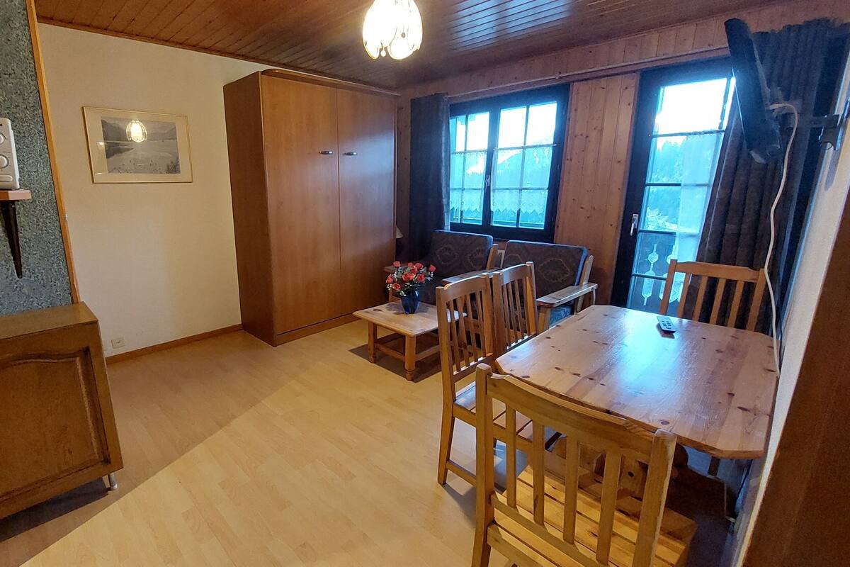 A wooden dining table with chairs, a small side table with a vase, a microwave on a shelf, a wardrobe, and a window with curtains.