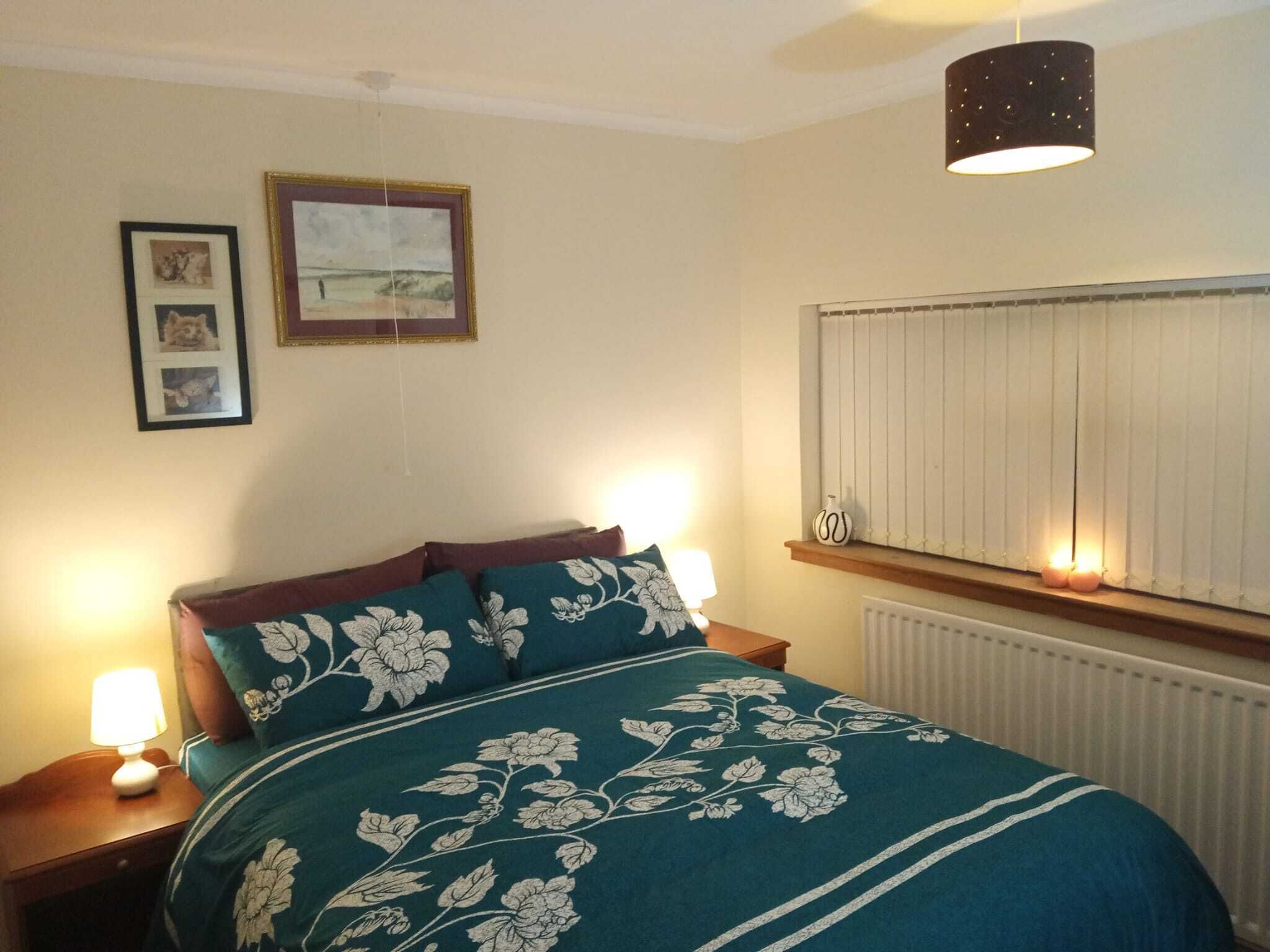 A bedroom with a bed, two bedside lamps, a window with blinds, and a framed picture on the wall.
