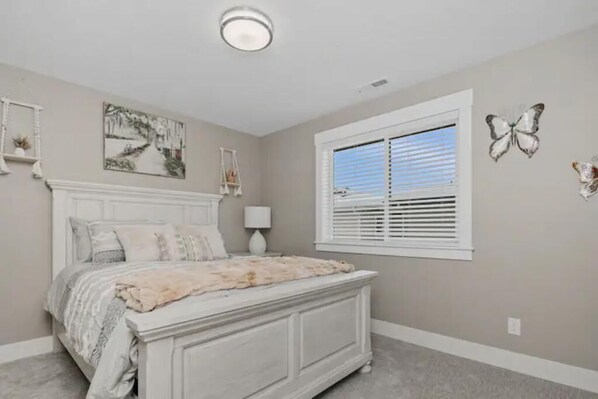 A bedroom with a white bed, a window with blinds, a wall-mounted lamp, and a painting.