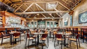 Un restaurante espacioso con techos altos y vigas a la vista, que cuenta con una zona de bar con estantes llenos de botellas y diversas opciones de asientos, incluyendo mesas y sillas.