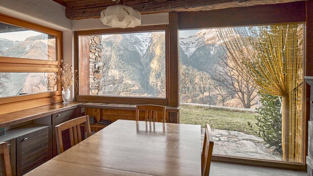 A rustic kitchen with wooden cabinets, a large wooden table, and a view of snow-capped mountains through large windows. - Secluded Mountain Gem: 1800m² Private Garden, Outdoor Kitchen & Stunning Views (Faido)