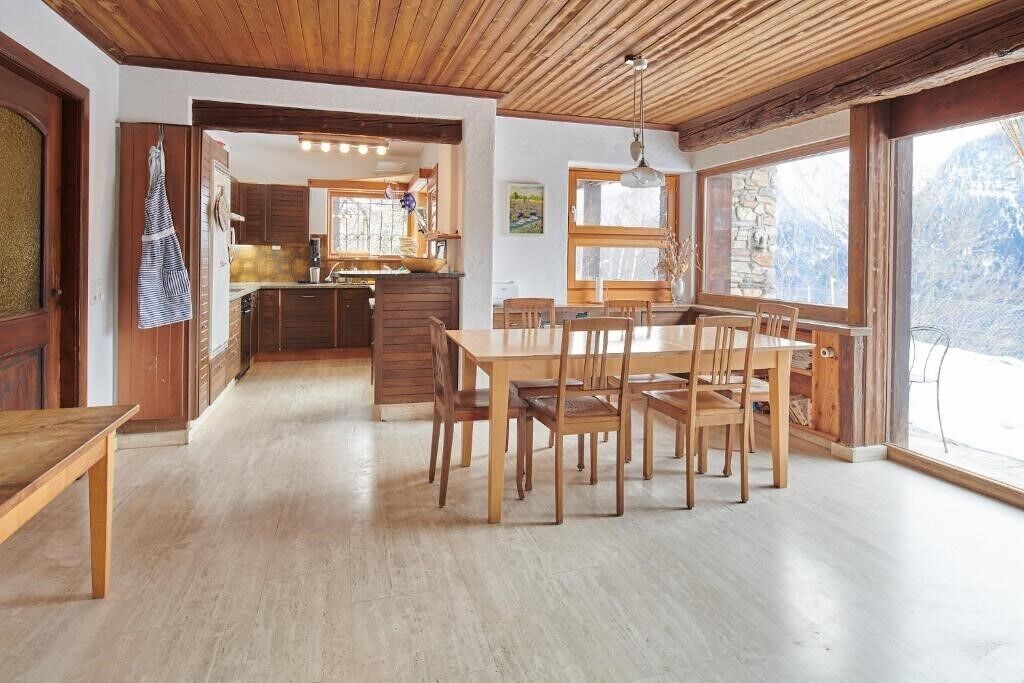 Een houten eettafel met stoelen, een keuken met houten kastjes en een groot raam met uitzicht op een besneeerd landschap.