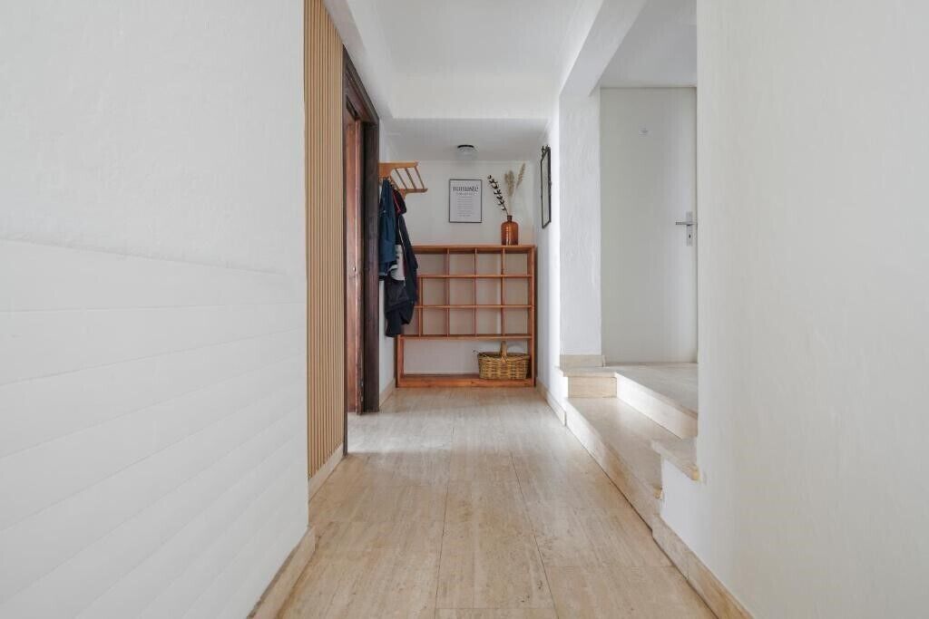 Een smalle gang met houten vloer, een houten bankje met een mand en een deur aan het einde.