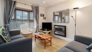 A living room with a sofa, armchair, TV, fireplace, and a table with a bowl and glasses. - Park Place Court 2 - Close to Beach, Parking (Tenby)