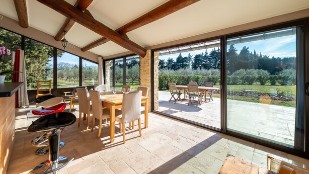 A spacious dining area with wooden tables and chairs, large windows offering a view of the outdoors, and a stone floor. - Gîte du Mas Saint Paul - Maison avec piscine (Travaillan)