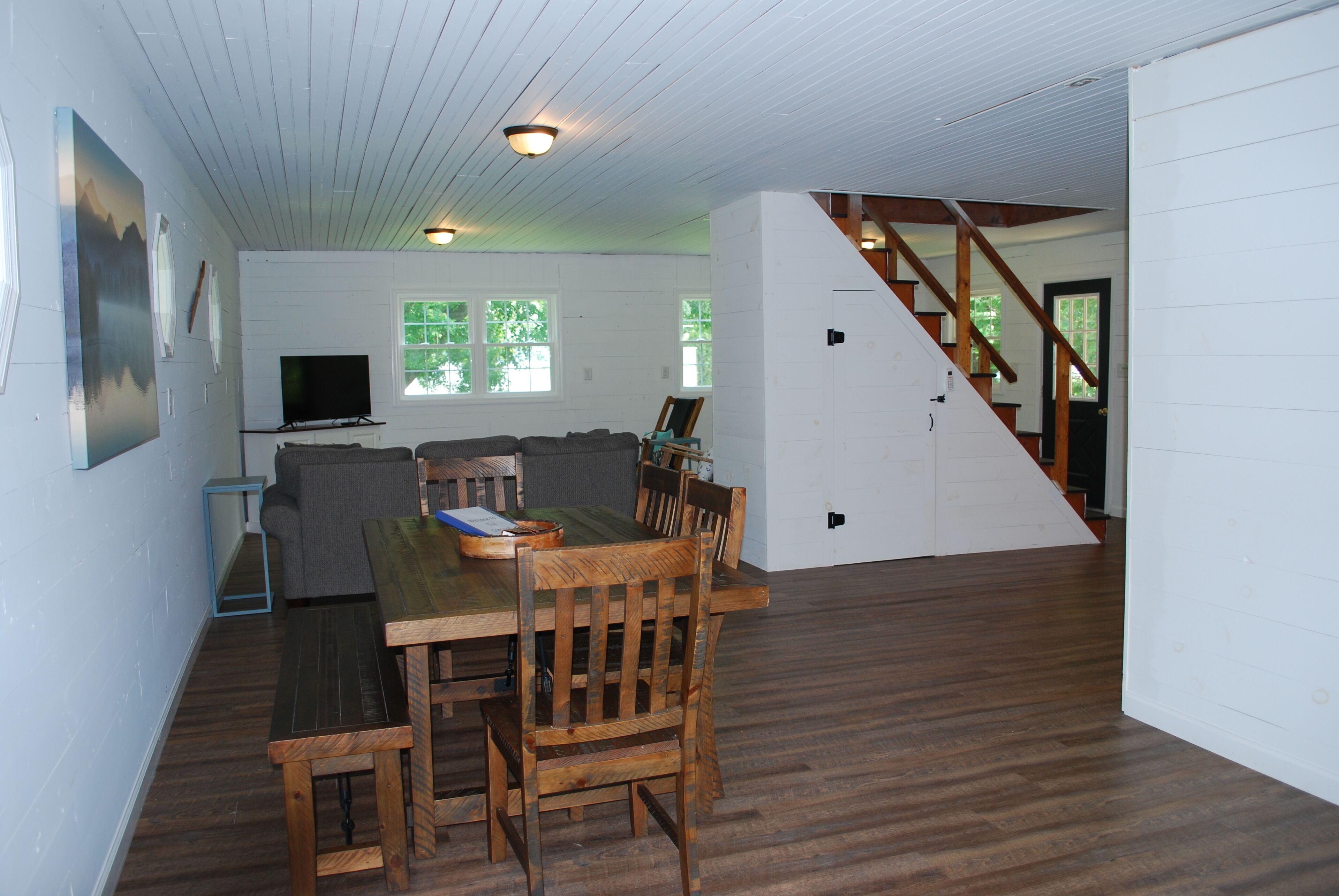 A spacious living area with a wooden dining table and chairs, a sofa, and a television. A staircase leads to an upper level.
