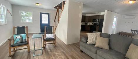 A spacious living room with a grey sofa, two wooden chairs, and a staircase leading to a kitchen.